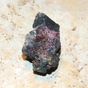 May include: A dark gray mineral specimen with patches of pink and red crystal formations. The rock has a rough texture and a mix of colors, including black, gray, and white. The specimen is a natural, unpolished stone.