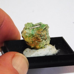 May include: A close-up of a mineral specimen. The rock has a rough texture with green, orange, and brown hues. A small, silver-colored metallic piece is embedded in the rock. The specimen is displayed on a black square base.