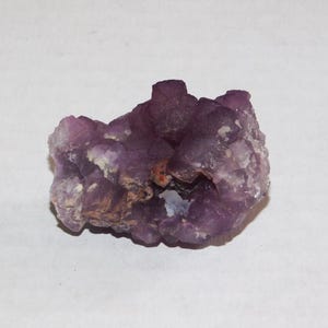 May include: A cluster of amethyst crystals, showcasing a deep purple hue with lighter shades in the crevices. The rough, natural texture of the stone is visible, with small crystalline formations. The overall shape is irregular and organic.
