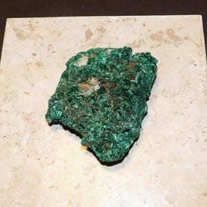 Puede incluir: Un espécimen mineral de malaquita verde vibrante con una estructura cristalina. El mineral se encuentra sobre una baldosa cuadrada de color beige claro, mostrando su rico color y textura. La superficie de la malaquita tiene un aspecto rugoso e irregular.