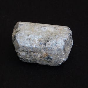 May include: A light grey mineral specimen, rectangular in shape, with a textured surface. The rock has silver highlights and is set against a black backdrop. The stone appears to be natural and unpolished.