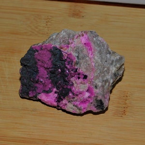 May include: A pink and black mineral specimen with a rough, irregular shape. The mineral is embedded in a grey rock matrix.