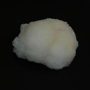 Puede incluir: Un espécimen mineral claro y texturizado sobre un fondo negro. El mineral tiene una forma redondeada con una superficie ligeramente rugosa y un tono crema pálido. La apariencia general sugiere una formación geológica natural.
