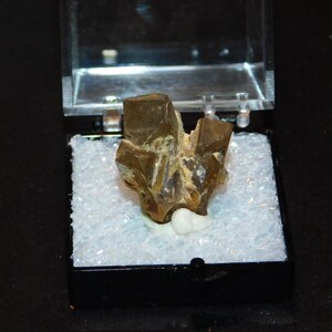 May include: A brown mineral specimen with a crystalline structure, presented in a clear plastic case with a black base. The mineral is mounted on a white base. The case is open, revealing the mineral's details.