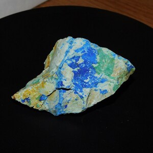 May include: A rough, irregular shaped rock with patches of blue, green, and white. The rock is on a black surface.