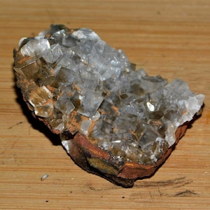 May include: A cluster of clear, cubic crystals with a slight brown tint, set on a reddish-brown matrix. The crystals are densely packed, creating a textured surface. The mineral specimen is displayed on a light brown wooden surface.