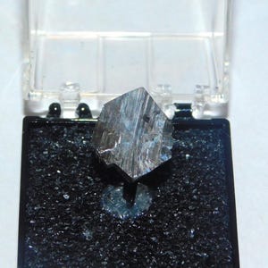 May include: A metallic, cube-shaped mineral specimen, possibly pyrite, displayed in a clear plastic case with a black base. The mineral has a reflective, silver-gray surface and sits on a bed of small, dark granules.