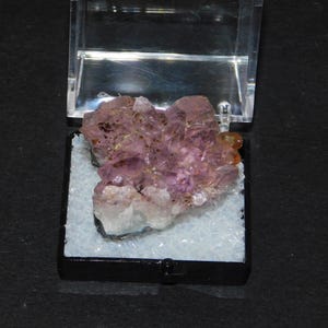 Puede incluir: Un grupo de cristales de amatista, que muestra una gama de tonos púrpuras, se presenta en una vitrina transparente con bisagras. Los cristales están colocados sobre un lecho de material granular blanco, sobre un fondo negro. También es visible un pequeño cristal de color naranja.