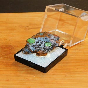 May include: A small, dark green and brown rock with two bright green crystals, displayed in a clear plastic box on a wooden surface.