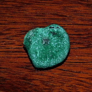 Puede incluir: Una piedra de malaquita verde con un patrón radial y un centro gris oscuro. La piedra es lisa y tiene un acabado brillante, casi metálico.