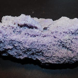 Puede incluir: Un espécimen mineral cristalino en bruto que presenta una estructura densa y porosa con un tono violeta lavanda claro. La superficie está cubierta de diminutas puntas de cristal brillantes que crean una textura rugosa e irregular.