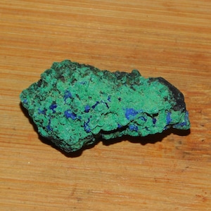 Puede incluir: Un espécimen mineral de malaquita y azurita verde y azul áspero. El mineral tiene una superficie áspera y desigual y es de color verde oscuro con acentos azules.