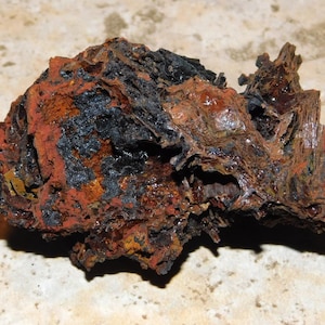 May include: A close-up of a rough, irregularly shaped rock with a mix of colors. The rock has a dark, almost black, surface in some areas, with patches of rusty orange and brown. The texture appears coarse and weathered.