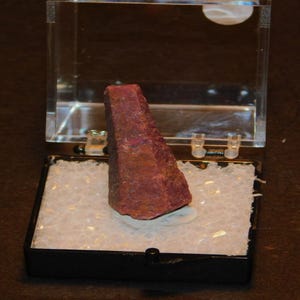 以下が含まれることがあります： 透明なプラスチック製のディスプレイケースに入った、濃い赤色の尖った粗削りされた宝石