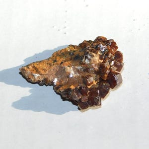 May include: A cluster of dark reddish-brown crystals on a rough, tan-colored matrix. The crystals have a sparkling, reflective quality, and the overall appearance suggests a natural mineral specimen.