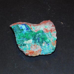 Pode incluir: Um espécime mineral colorido com uma superfície áspera e texturizada. A rocha apresenta uma mistura de cores vibrantes, incluindo tons de verde, azul e vermelho. A pedra natural tem formato irregular e parece ser uma peça bruta e não polida.