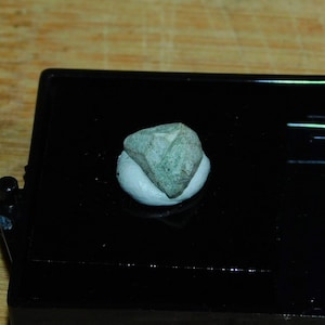 Puede incluir: Una piedra verde facetada, similar a un cristal, sobre un fondo blanco. La piedra está en una caja de presentación negra.