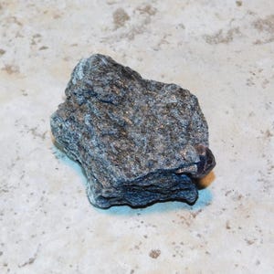 Może przedstawiać: Ciemnoszara skała o warstwowej fakturze i srebrnych plamkach. Skała ma nieregularny kształt i wydaje się być okazem mineralnym. Powierzchnia ma małe, odblaskowe plamki, prawdopodobnie piryt. Spoczywa na jasnej, nakrapianej powierzchni.