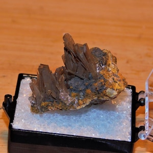 Puede incluir: Un cúmulo de cristales marrones translúcidos sobre una roca gris oscura. Los cristales están dispuestos en forma de abanico y tienen una apariencia puntiaguda y afilada. La roca está rodeada de un material blanco y espumoso, que probablemente sea un tipo de material de embalaje. La roca y los cristales se muestran en una caja negra con una tapa de plástico transparente.