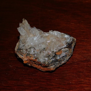 Puede incluir: Un grupo de cristales transparentes y translúcidos sobre una roca rugosa, marrón y gris. Los cristales son afilados y puntiagudos, y están dispuestos en un patrón aleatorio.