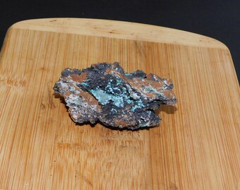 Può includere: Un sasso ruvido di colore grigio scuro con una macchia di cristalli blu turchese e bianchi. Il sasso è appoggiato su una superficie di legno.