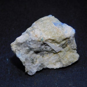 Puede incluir: Una roca rugosa de color gris claro con un pequeño cristal blanco triangular incrustado. También hay pequeños cristales azules dispersos por toda la roca.