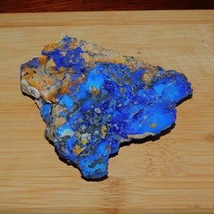 May include: A vibrant blue mineral specimen with patches of tan and gray. The stone has a rough, uneven texture and is displayed on a wooden cutting board. The mineral's striking blue color is the focal point.