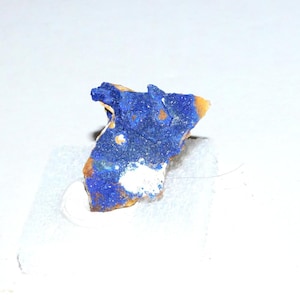 May include: A small, irregularly shaped mineral specimen with a vibrant blue hue, accented by patches of white and hints of orange-brown. The mineral is mounted on a clear adhesive, showcasing its natural texture and color variations.