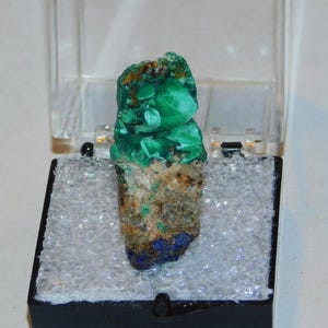 Könnte beinhalten: Ein lebhaft grünes Malachit-Mineralexemplar, präsentiert in einer transparenten Kunststoffbox mit schwarzem Sockel. Der Malachit weist Schichten aus grünen, braunen und blauen Farbtönen auf, die seine natürlichen Muster hervorheben. Die Box ist geöffnet.