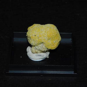 Può includere: Un campione di minerale giallo con una superficie ruvida e strutturata, esposto su un vassoio nero. Il minerale ha una forma arrotondata e poggia su una base bianca. Lo sfondo è nero pieno.