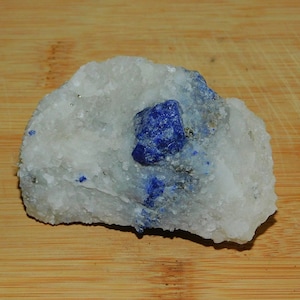 May include: A blue Lapis Lazuli crystal embedded in a white matrix of rock. The crystal has a sharp, geometric shape and is surrounded by smaller, blue crystals.