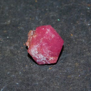 Puede incluir: Un espécimen de cristal hexagonal en bruto con un tono rosa intenso y vibrante. La piedra preciosa presenta facetas naturales sin pulir y una superficie ligeramente texturizada con inclusiones minerales blancas opacas visibles.