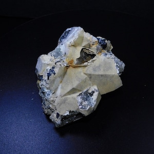 Puede incluir: Un espécimen mineral natural en bruto con una estructura geométrica compleja. La piedra presenta una mezcla de tonos beige, gris y gris azulado oscuro. La superficie tiene una apariencia cristalina con facetas e inclusiones visibles, sobre un fondo oscuro.