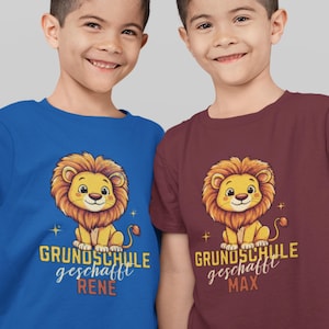 Könnte beinhalten: Zwei Kinder tragen blaue und weinrote T-Shirts mit einem Comic-Löwen-Motiv. Die Shirts sagen "Grundschule geschafft" mit den Namen "Rene" und "Max" darunter.