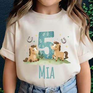 Peut inclure: T-shirt crème avec un motif d'anniversaire. Il présente deux chevaux de dessin animé, le chiffre 5 en bleu sarcelle avec des détails floraux, des fers à cheval et le nom "MIA" en bleu sarcelle. Vêtement pour enfants.