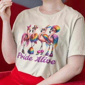 Könnte beinhalten: Cremefarbenes T-Shirt mit einer farbenfrohen Illustration von vier Flamingos, die mit regenbogenfarbenen Federn, Accessoires und Sonnenbrillen geschmückt sind. Der Text "Pride Alive" ist in lila Schrift geschrieben. Das T-Shirt wird von einer Person getragen.