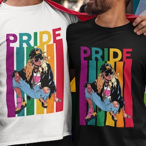 Könnte beinhalten: Zwei T-Shirts, eines weiß und eines schwarz, jeweils mit einer farbenfrohen Grafik. Die Grafik zeigt das Wort "PRIDE" in Regenbogenfarben über einer Illustration einer Person mit Mütze, Sonnenbrille und zerrissenen Jeans. Die T-Shirts werden von zwei Personen getragen.