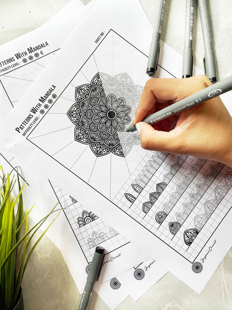 May include: A hand using a grey Staedtler pigment liner 0.1 pen to colour a black and white mandala design on white paper. The paper is labelled "Patterns With Mandala" and "Difficulty Level". Several other mandala design sheets and pens are scattered around.
