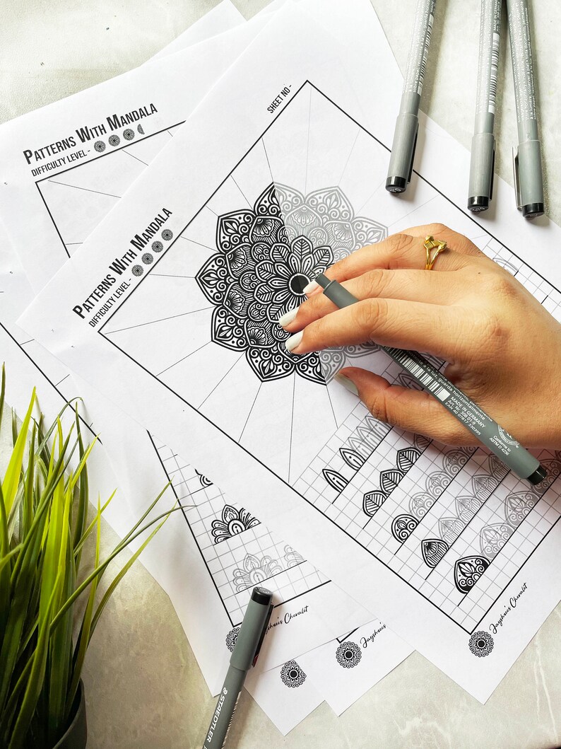 May include: A collection of mandala coloring pages with the text "Patterns With Mandala" and "Difficulty Level". A hand is holding a grey pen, coloring a mandala design. Grey pens are scattered around the pages.