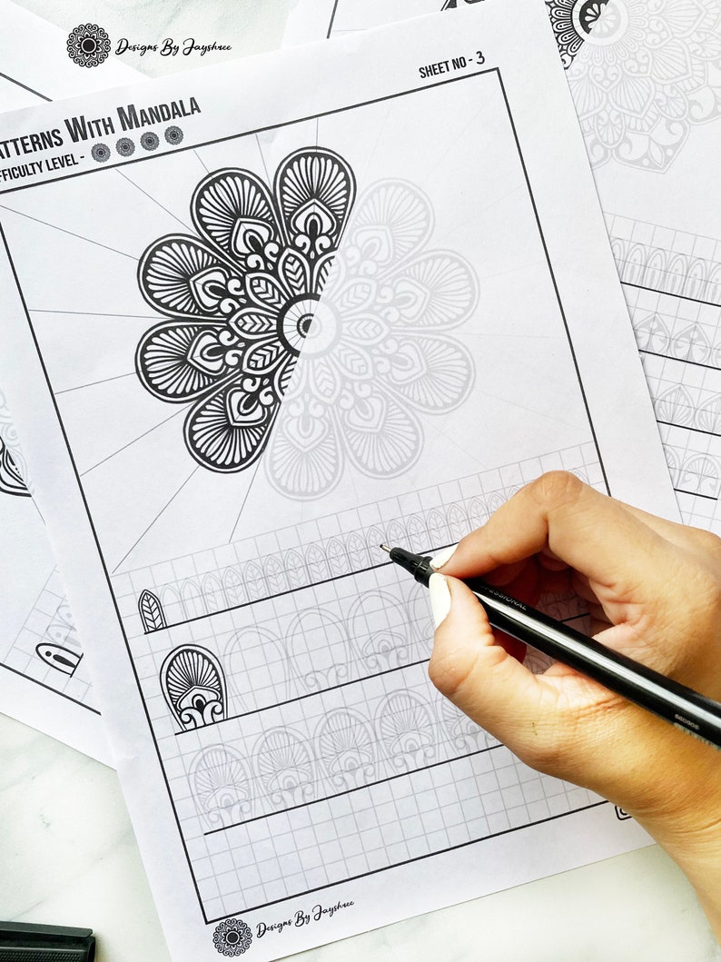 May include: Black and white mandala pattern sheets with the text "PATTERNS WITH MANDALA" and "SHEET NO-3". A hand is holding a black pen, drawing on the sheet. The sheets have a grid pattern and mandala designs.