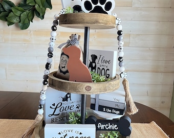 May include: A three-tiered wooden tiered tray decorated with dog-themed decorations. The top tier features a white birdhouse with a keyhole, a black paw print, and a small potted plant. The middle tier features a brown burlap bag with the word "dog" on it, a brown dog figurine, and a white sign with the text "I love my dog". The bottom tier features a white sign with the text "You can't buy love but you can rescue", a white sign with the text "Love is wet noses and wagging tails", and a black bone-shaped sign with the text "Archie".