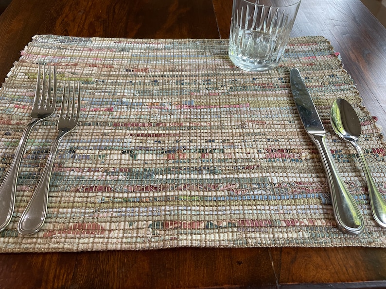 May include: A place setting with two forks, a knife, and a spoon on a woven placemat with a light brown background and multi-colored stripes. A clear glass is on the table to the left of the place setting.