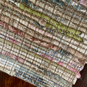 May include: A close-up of a woven rug with a beige background and colorful stripes of pink, green, and blue. The rug is made of recycled fabric and has a textured surface.