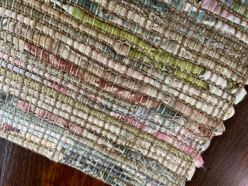 May include: A close-up of a woven rug with a beige background and colorful stripes of pink, green, and blue. The rug is made of recycled fabric and has a textured surface.