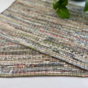 May include: Two woven placemats in a neutral color palette with a variety of textures. The placemats are made of recycled materials and have a rustic, handmade look.