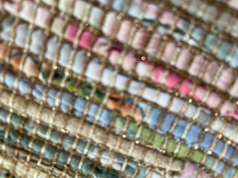 May include: Close-up of a woven fabric with a textured, multi-colored pattern. The fabric is made of a variety of materials, including natural fibers and metallic threads. The colors are muted and earthy, with shades of beige, brown, pink, blue, and green.