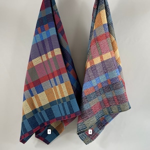 May include: Two colourful woven kitchen towels with a plaid pattern in shades of red, blue, yellow, green, and purple. The towels are hanging on a white wall.