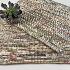May include: A woven rug made from recycled materials in shades of brown, beige, green, and pink. The rug is made of strips of fabric woven together to create a textured surface.