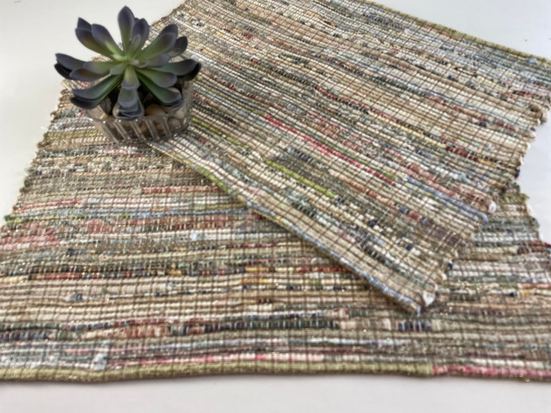 May include: A woven rug made from recycled materials in shades of brown, beige, green, and pink. The rug is made of strips of fabric woven together to create a textured surface.