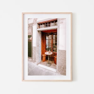 May include: A small, round table with two chairs sits outside a cafe entrance in a narrow alleyway. The cafe is called "Caffe Ottie" and the building has a red door and a tiled wall.